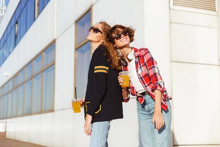 Two joyful teenage girls in sunglasses drinking orange juice while standing outdoorsの写真素材