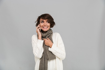 Image of emotional young lady wearing scarf standing isolated over grey background. Looking aside talking by mobile phone.の写真素材