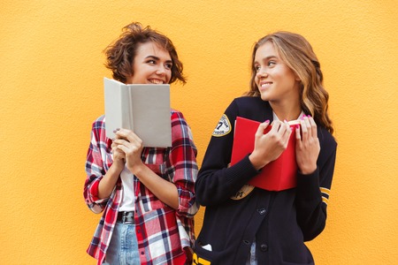 Portrait of two happy pretty teenage girls with books standing and looking at each other over yellow backgroundの写真素材