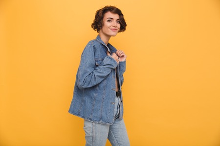 Portrait of a young smiling woman dressed in denim jacket posing while standing and looking at camera isolated over yellow backgroundの写真素材