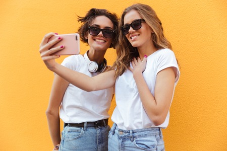 Two cheery young teenage girls in sunglasses taking a selfie while standing against orange wall