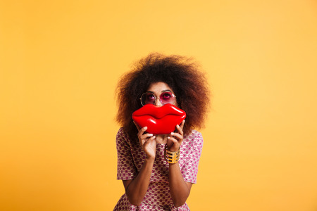 Pretty retro stylish african woman having fun while posing with big red lips, looking upward, isolated on yellow backgroundの写真素材