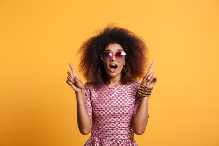 Close-up portrait of retro stylish amazed african wooman in sunglasses pointing with two fingers up, looking upward, isolated on yellow backgroundの写真素材