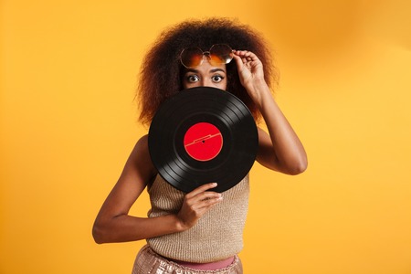 Portrait of a pretty afro american woman dressed in retro clothes posing while holding vinyl isolated over yellow backgroundの写真素材