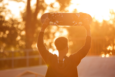 Back view of a young male teenager guy holding skateboard on shoulders and looking at sunset outdoorsの写真素材