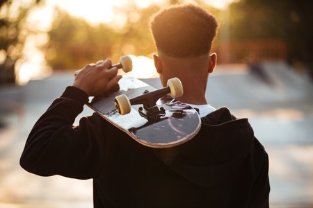 Back view of a male teenager guy holding skateboard on shoulders while standing outdoorsの写真素材