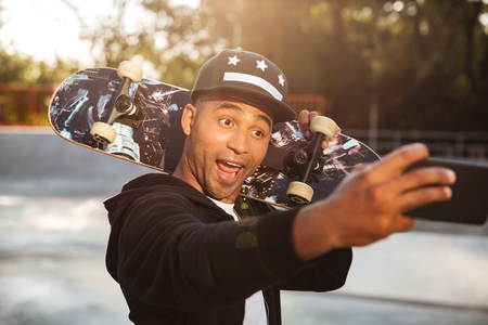 Portrait of a joyful african male teenager taking a selfie while holding a skateboard on his shoulders outdoorsの写真素材