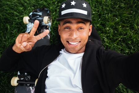 Top view of a happy african male teenager lying on a grass with a skateboard and taking a selfie outdoorsの写真素材