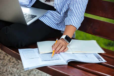 Close up of a female hand holding pencil above textbook on the bench with laptop outdoorsの写真素材