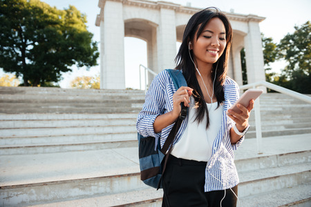 Young asian woman in striped shirt checking her phone, while going down stairsの写真素材
