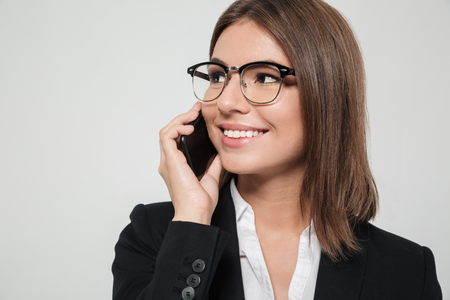 Close up portrait of a young pleased businesswoman in eyeglasses talking on mobile phone and looking away isolated over white backgroundの写真素材