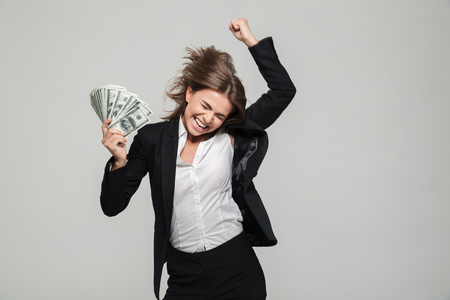 Portrait of a thrilled excited businesswoman in suit holding bunch of money banknotes and celebrating isolated over white backgroundの写真素材