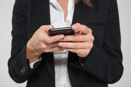 Close up portrait of a young woman in suit typing message on mobile phone isolated over white backgroundの写真素材