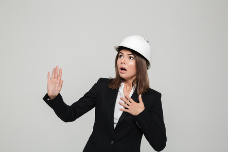 Portrait of a young scared woman in suit and hard hat looking away and gesturing with hands isolated over white backgroundの写真素材
