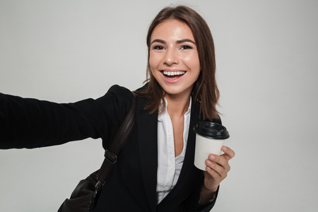 Portrait of a smiling attractive woman in suit taking a selfie while holding take away coffee cup isolated over white backgroundの写真素材