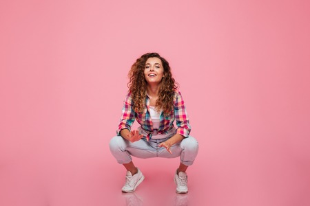 Young funny lady in casual clothes squatting and smiling to camera isolated over pinkの写真素材