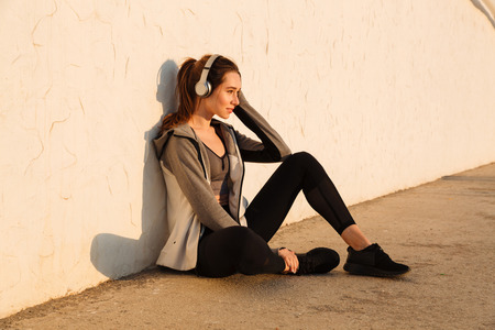 Portrait of cheerful young sport woman listening to music, resting after workout, sitting near white wallの写真素材