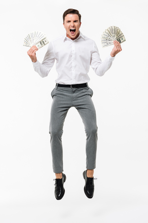 Full length portrait of a happy successful man in white shirt holding bunch of money banknotes while jumping and looking at camera isolated over white backgroundの写真素材