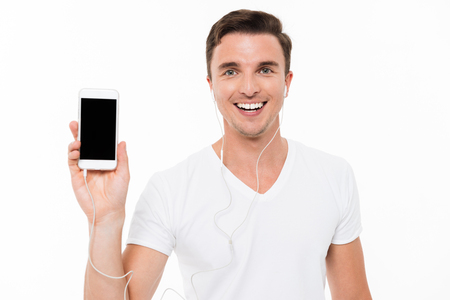 Portrait of a cheerful confident man in earphones pointing finger at blank screen mobile phone while standing and looking at camera isolated over white backgroundの写真素材