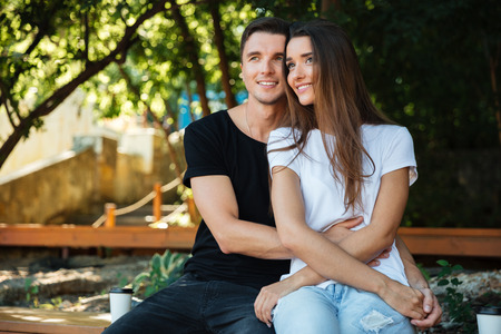 Portrait of a smiling attractive couple in love sitting on the bench and hugging at the park outdoorsの写真素材