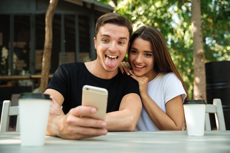 Portrait of a funny young couple taking selfie while sitting together at the cafe outdoorsの写真素材