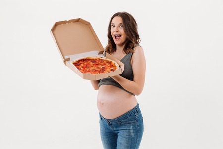 Photo of surprised pregnant woman standing isolated over white background. Looking camera holding pizza.の写真素材