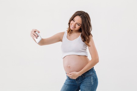 Photo of happy pregnant woman isolated over white background make selfie with her belly by mobile phone. Looking aside.の写真素材