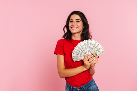 Smiling brunette woman holding money in hands and looking at the camera over pink backgroundの写真素材