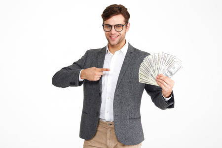 Portrait of pleased young man in eyeglasses and a jacket holding bunch of money banknotes while standing and pointing finger isolated over white backgroundの写真素材