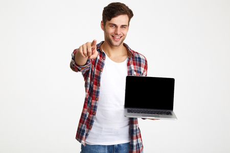 Portrait of a confident young man holding blank screen laptop computer and pointing finger at camera isolated over white backgroundの写真素材