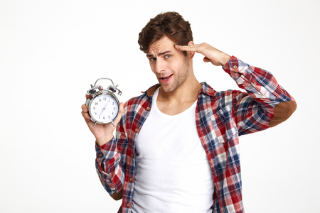 Portrait of a handsome casual man with hand at his head holding alarm clock isolated over white backgroundの写真素材