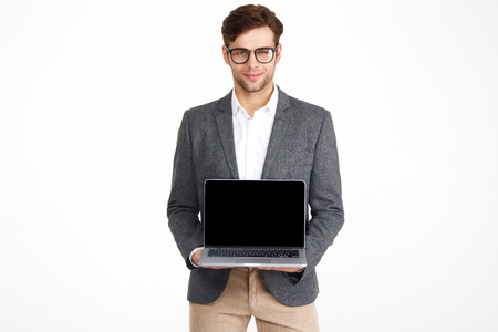 Portrait of a young smiling businessman in eyeglasses and a jacket showing blank screen laptop computer while standing and looking at camera isolated over white backgroundの写真素材