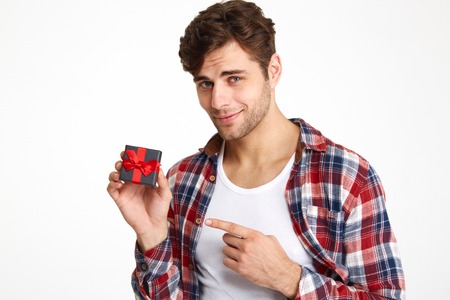 Portrait of a charming brunette man pointing finger at a little present box and looking at camera isolated over white backgroundの写真素材