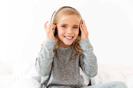 Close-up of pretty smiling little girl touching her headphones while listening to music, looking at cameraの写真素材
