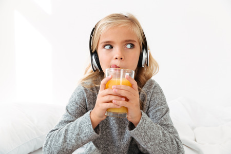Close-up photo of pretty little girl in headphones licking while drinking orange juice, looking asideの写真素材