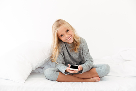 Cheerful girl in gray pajamas holding smartphone, looking at camera while sitting on bedの写真素材