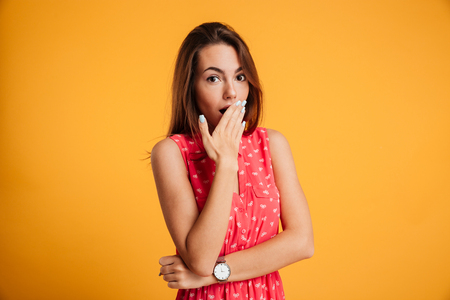 Photo of surprised pretty girl in red dress standing with her hand covering mouth and looking at camera isolated on yellow backgroundの写真素材