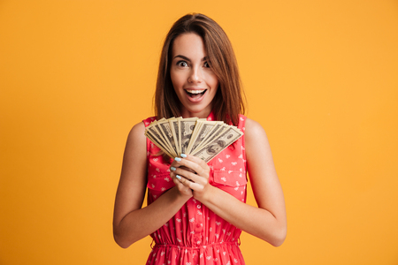 Happy young charming woman in red dress holding bunch of dollar bills, looking at camera, isolated on yellow backgroundの写真素材