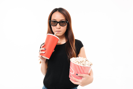 Portrait of a young asian girl in 3d glasses drinking from a cup with a straw while holding popcorn box and looking at camera isolated over white backgroundの写真素材