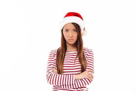 Portrait of an upset asian girl in christmas hat standing with arms folded isolated over white backgroundの写真素材