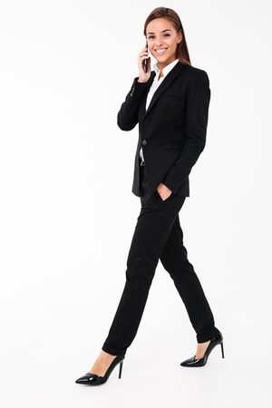 Photo of cheerful pretty businesswoman walking isolated over white background talking by phone holding folders. Looking camera.の写真素材