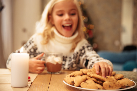 Blurred photo of happy little girl taking sweet cookie while sitting at festive table, selective focus on handの写真素材