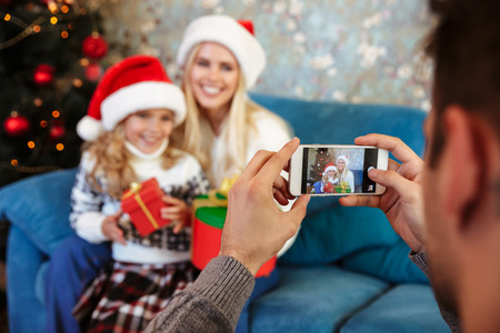Cropped view of father taking photo on mobile phone of his wife and daughter in Santa's hat, selective focus on phoneの写真素材