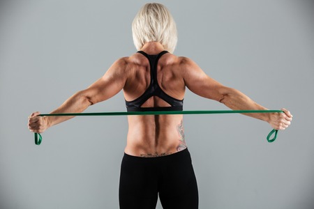 Back view portrait of a strong muscular adult sportswoman working out with a stretching band...
