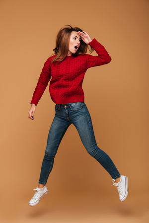 Full length portrait of amazed pretty brunette woman in red knitted sweater jumping over beige background, looking asideの写真素材
