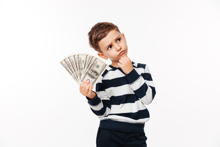 Portrait of a thoughtful cute little kid showing bunch of money banknotes and looking away at copy space isolated over white backgroundの写真素材