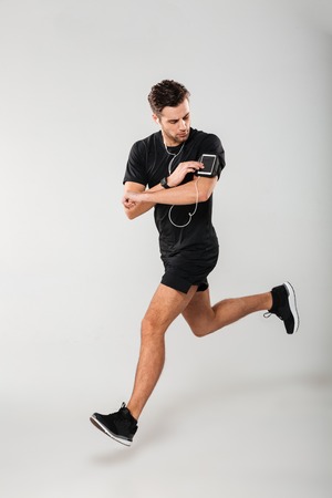 Full length portrait of a handsome young man athlete in earphones listening to music while running and touching his phone in armband isolated over gray backgroundの写真素材