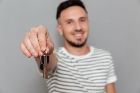 Smiling man in t-shirt showing keys at the camera over gray background. Focus on keysの写真素材