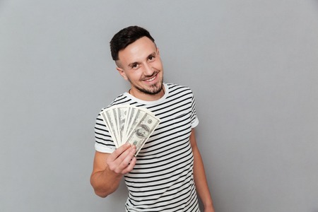 Smiling man in t-shirt holding money and looking at the camera over gray backgroundの写真素材