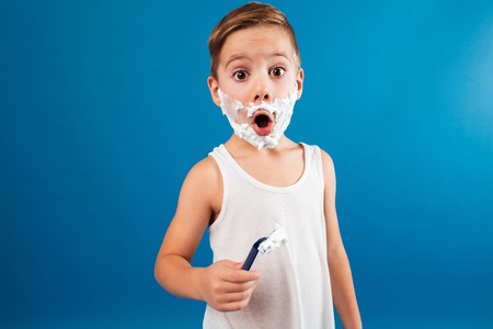 Shocked young boy in shaving foam like man holding razor and looking at the camera over blue backgroundの写真素材